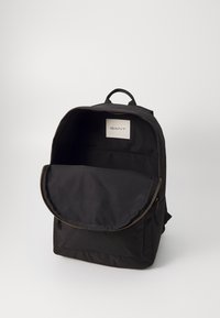 Sac à dos en tissu noir avec une fermeture éclair, un rabat avant et une poche intérieure. Présente une étiquette avec le logo "GANT". Design simple et fonctionnel.