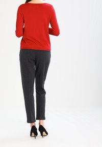 Femme portant un haut à manches longues rouge, un pantalon gris foncé à motif et des chaussures à talons noirs, debout de dos.