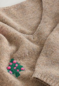 Pull en laine beige avec col en V et broderie florale texturée verte et rose sur le devant, près de l'ourlet.