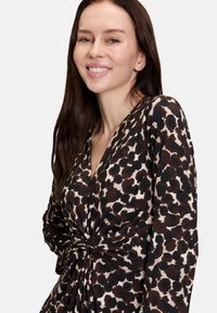 Femme aux longs cheveux bruns souriante, portant une robe à manches longues avec un motif abstrait à taches noires et brunes et un détail de nœud froncé.