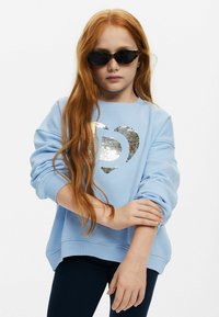 Sudadera azul claro con un diseño de corazón de lentejuelas, puños y dobladillo acanalados, combinada con gafas de sol negras. Larga melena castaña cae sobre los hombros.