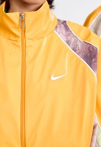 Žuta jakna na zatvarač s visokim ovratnikom, s sjajnom završnom obradom, apstraktnim tkaninskim detaljima na ramenima i bijelim Nike logom.