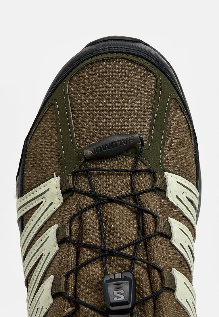 Brązowe tekstylne buty sportowe z zielonymi akcentami, gruba czarna gumowa podeszwa i skomplikowany system sznurowania. Posiadają logo Salomon na górze.