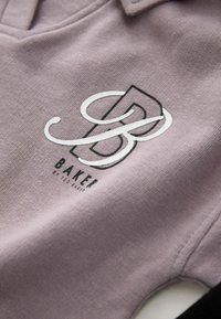 Lyse lilla, tekstureret poloshirt med en sort og hvid logo med teksten "BAKEI". Indeholder klassisk krave og syede detaljer.