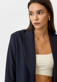 Marineblauwe gestreepte blazer met scherpe revers, gedragen over een crèmekleurige crop top. Glad materiaal, losse pasvorm, minimale hardware.