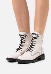 Bottes courtes en cuir blanc avec lacets noirs, bout rond et semelle noire texturée avec un petit talon bloc et une couture contrastée.