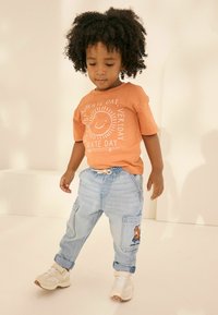 Orange T-shirt with white sun and text, paired with light blue denim joggers featuring side pockets and cartoon design, finished with white sneakers.