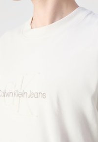 Bílý bavlněný tričko s kulatým výstřihem, na hrudi s vyšitým logem "CK" a "Calvin Klein Jeans" v světlé béžové barvě.