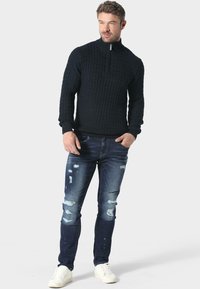 Czarny sweter robiony na drutach z zamkiem na szyi, z prążkowanymi mankietami, zestawiony z dopasowanymi, przetartymi niebieskimi dżinsami i białymi trampkami z lekkimi zarysowaniami.