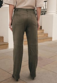 Pantalon taille haute en daim doux vert olive, jambes droites, poches arrière et texture lisse, porté avec des talons aiguilles.