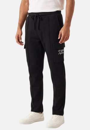 Hombre con pantalones cargo negros con cintura de cordón y zapatillas blancas, recortado a la mitad del torso sobre un fondo liso.