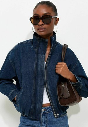 Woman wearing large black sunglasses and hoop earrings, dressed in a dark blue denim jacket and jeans, holding a dark brown handbag.