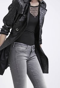 Abrigo negro perforado con una superficie brillante, superpuesto a una blusa negra con detalles de malla, combinado con unos jeans ajustados grises.