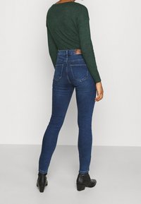 Mörkblå högmidjade skinny jeans i denim med två bakfickor och en logopatch bak; matchas med en långärmad grön topp.
