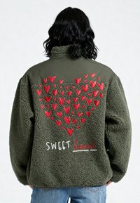 Persona che indossa una giacca in pile verde con disegni di cuori rossi e la scritta "SWEET heart" ricamata sulla schiena, abbinata a jeans blu.