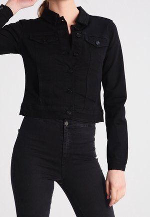 Vrouw draagt een getailleerd zwart denim jack met knopen aan de voorkant en bijpassende zwarte jeans met hoge taille tegen een witte achtergrond.