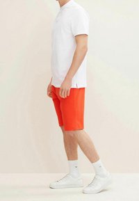 Biała koszulka polo z krótkim rękawem, pomarańczowe szorty i białe sneakersy. Koszulka polo ma zapięcie na guziki przy kołnierzyku; szorty są do kolan i gładkie.