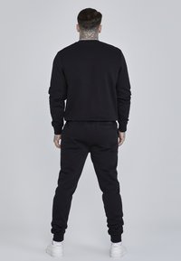 Zwarte sweatshirt en sweatpants combinatie, gemaakt van zacht materiaal, met ribgebreide manchetten en een comfortabele pasvorm. Witte sneakers maken de look compleet.