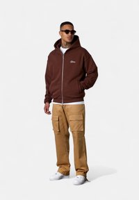 Braune Zip-Hoodie mit einer Kordelzugkapuze, kombiniert mit beigen Cargo-Hosen mit mehreren Taschen. Das Modell trägt weiße Sneaker und Sonnenbrillen.