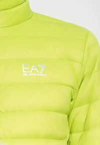 Erks-roheline puhvkattega jakk, millel on õmmeldud paneelid, kõrge krae ja valge EA7 Emporio Armani logo rinnal.