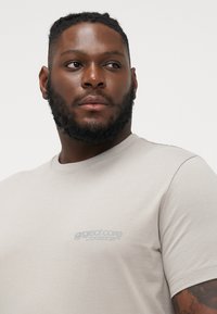 Lys beige t-skjorte laget av myk bomullsblanding, med rund hals og frontlogo i grått. Korte ermer og avslappet passform.