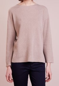 Pull en maille beige avec un col rond et des manches longues, présentant une coupe décontractée et des fentes latérales, associé à un pantalon sombre.