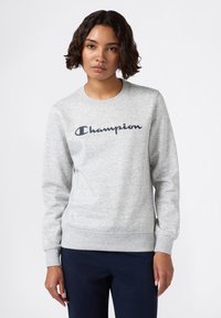 Vrouw met kort krullend haar in een lichtgrijze Champion sweatshirt en donkere broek, staande voor een effen lichte achtergrond.