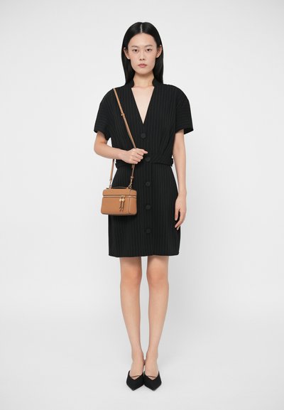 Γυναίκα φορώντας ένα μαύρο φόρεμα με κάθετες ρίγες, κοντό μανίκι και μεγάλα κουμπιά, μαύρες μυτερές γόβες και ένα καφετί τσάντα χιαστί, στέκεται μπροστά σε λευκό φόντο.