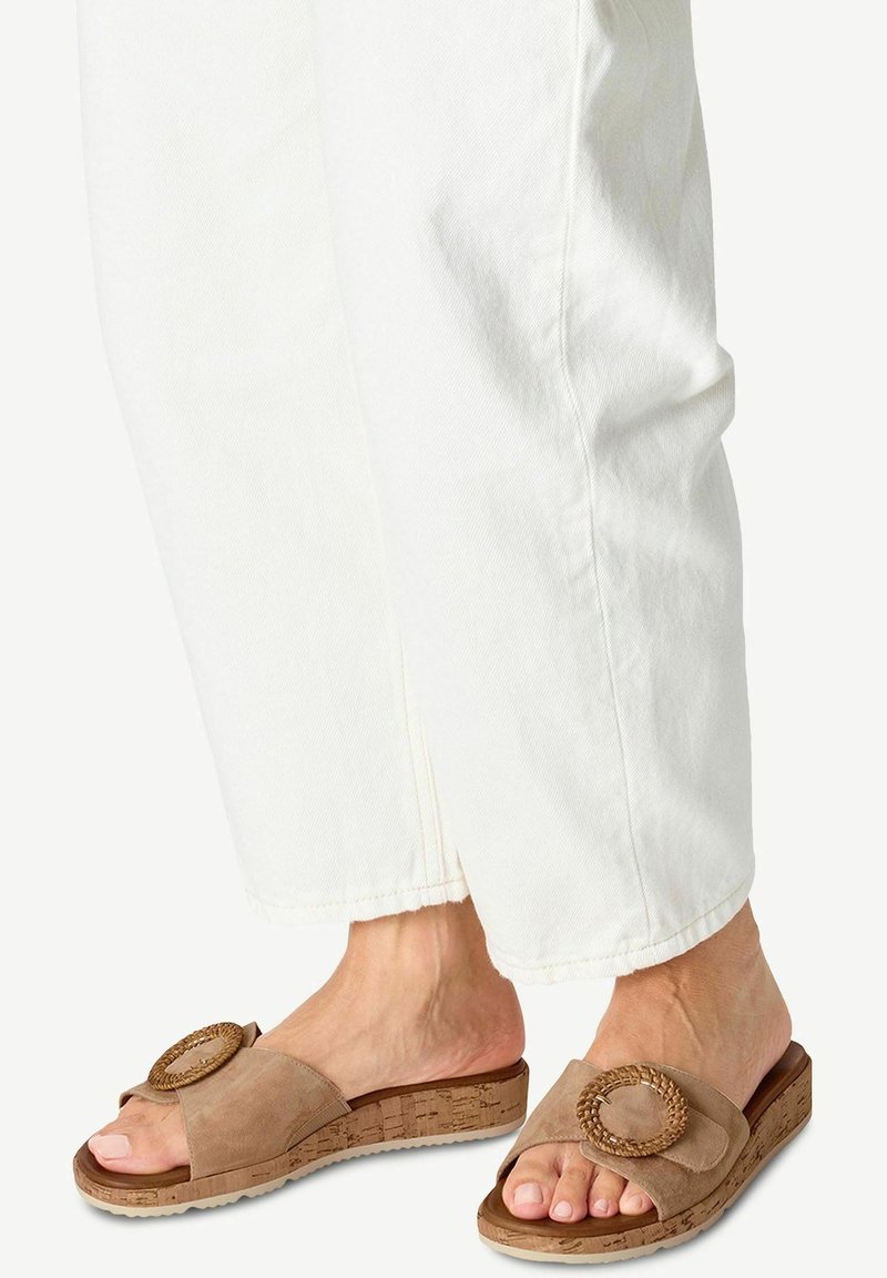 Sandales à glissière en suède tan avec accent circulaire tressé et semelle en liège ; portées avec un pantalon en denim blanc, mettant en valeur le design à bout ouvert.