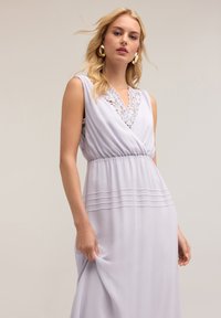 Helles lavendelfarbenes Maxikleid mit V-Ausschnitt, Spitzenakzenten und plissiertem Bund. Der Stoff ist glatt und hat eine fließende Silhouette.