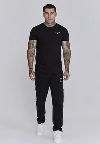 Svart T-shirt med korta ärmar och logotyp på bröstet, kombinerad med svarta cargobyxor med fickor och gulddetalj, avslutad med vita sneakers.