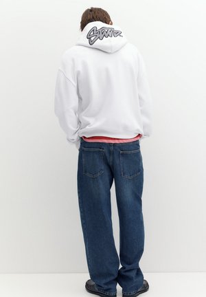 Persona che indossa una felpa bianca con cappuccio con scritta nera a graffiti sul cappuccio, jeans blu e scarpe nere, in piedi con le spalle rivolte verso uno sfondo bianco.