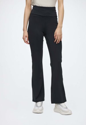 Donna che indossa pantaloni neri a vita alta con spacco all’orlo e sneakers bianche, in piedi davanti a uno sfondo chiaro e uniforme.