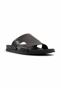 Sort åben tå slide sandal med bred læderrem, der har udsparinger på siden og et lille metalmærke på hvid baggrund.