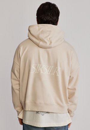 Man draagt beige capuchontrui met tekst "SIKSILK" op de achterkant, aan over een wit shirt en blauwe spijkerbroek, kijkt weg tegen een effen achtergrond.