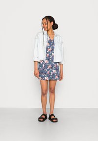 Vestido floral azul com mangas curtas, por baixo de uma leve jaqueta de ganga. Sandálias pretas completam o conjunto. Textura de tecido suave.