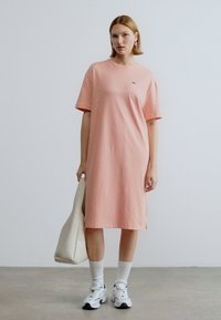 Ροζ βαμβακερό φόρεμα τύπου T-shirt με κοντά μανίκια, πλαϊνές σχισμές και μίνιμαλ λογότυπο. Συνδυάζεται με λευκές κάλτσες και αθλητικά παπούτσια, κρατώντας μια τσάντα tote.