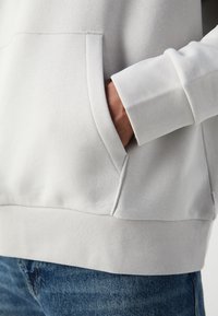 Hanorac gri deschis cu buzunar tip cangur, manșete și tiv din material ribbat. Prezintă o textură moale și un design minimalist. Asortat cu blugi albaștri.