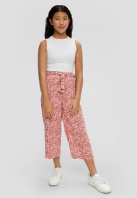 Mädchen mit langen dunklen Haaren, das ein weißes ärmelloses Top, pinkfarbene, blütenbedruckte, weite Hosen mit Gürtel und weiße Sneaker trägt und vor einem einfarbigen Hintergrund steht.