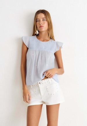 Femme portant un haut sans manches bleu clair avec des épaules volantées et un short blanc avec des détails cloutés, debout sur un fond blanc.