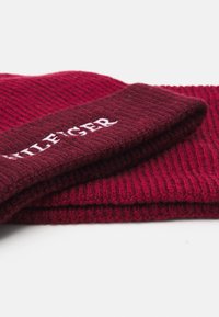 Röd ribbad stickad mössa med uppvikt kant och vitt broderat logotyp. Materialet verkar mjukt och strukturerat.