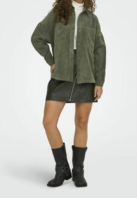 Kobieta ubrana w oversize'ową zieloną koszulę z korduroy, czarną skórzaną spódnicę z przednim zamkiem, czarne botki do połowy łydki oraz białą sweter z golfem.