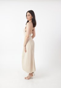 Lys beige, ærmeløs maxikjole med en tætsiddende silhuet, subtil tekstur og lav ryg, båret med strop-sandaler med høje hæle.