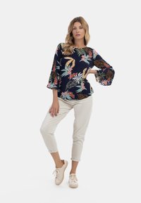 Blusa blu navale con stampa floreale, maniche lunghe a campana, abbinata a pantaloni cropped beige chiaro e sneakers crema. Tessuto morbido, vestibilità comoda.