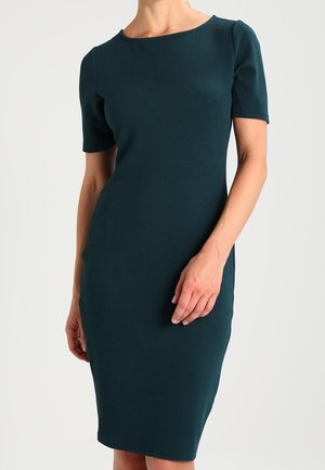 Femme portant une robe ajustée, longueur genou, manches courtes, de couleur bleu sarcelle avec un col rond, se tenant devant un fond clair uni.
