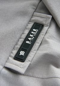 Harmaa kangas, jossa on tekstuuroitu pinta, ja siinä on suorakulmainen musta etiketti, jossa lukee "TB BAKER by TED BAKER" valkoisella letteringilla.