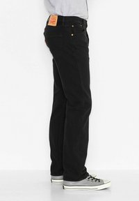 Svarta jeans i denim med rak passform, med en brun läderlapp på baksidan och ljusa sömnadsdetaljer. Kombinerade med grå sneakers.