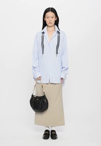 Črtasta modra oversize srajca z črnimi naramnicami, bež krilo, črna torbica in loafers čevlji. Model stoji na enobarvni podlagi.