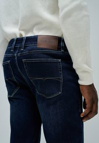 Vaqueros de mezclilla azul oscuro con un parche de cuero marrón con la marca "Salsa Jeans", costuras doradas y dos bolsillos traseros con solapas puntiagudas.