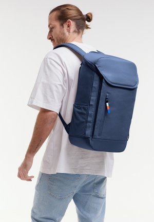 Man met teruggebonden haar, gekleed in een wit T-shirt en spijkerbroek, draagt een grote marineblauwe rugzak met meerdere vakken.